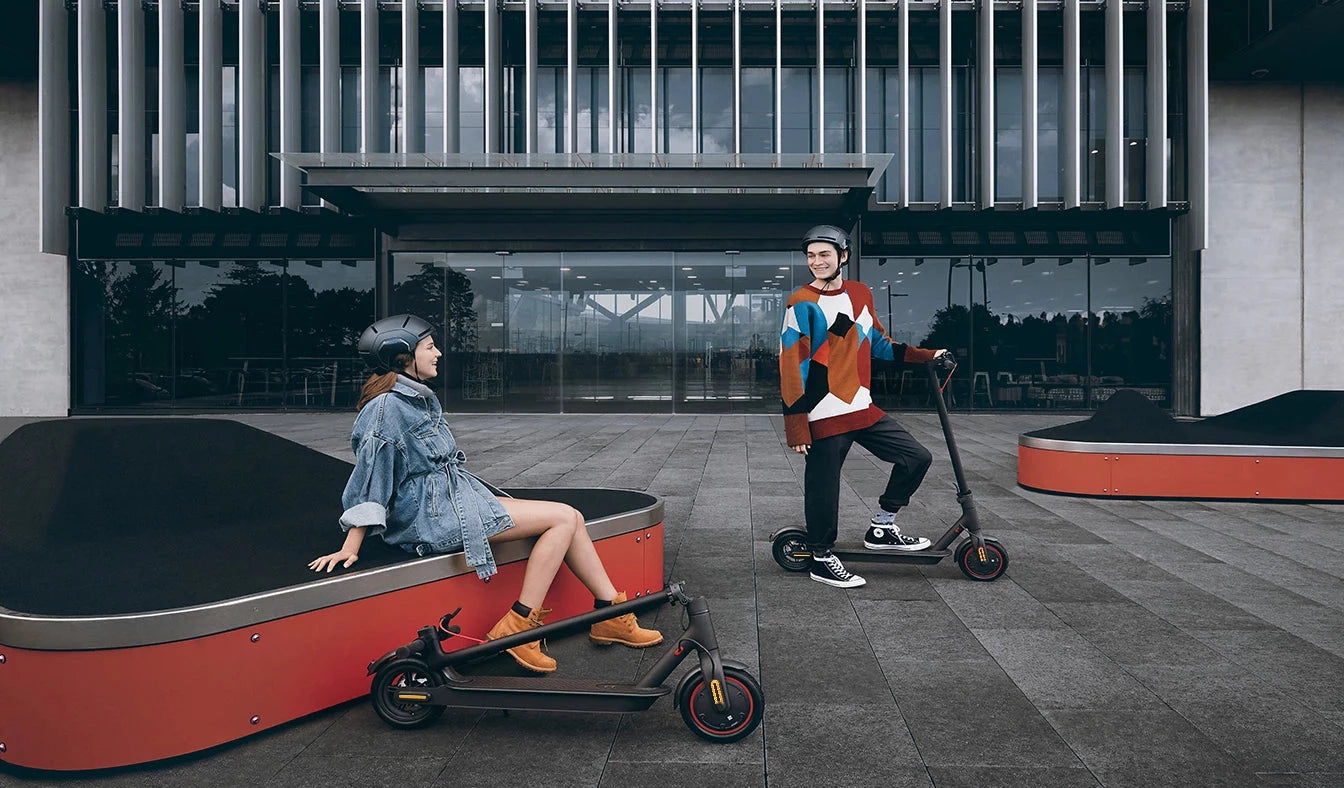 Lady and gentleman with helmets on their Xiaomi Esoocters outside a building on a bright day