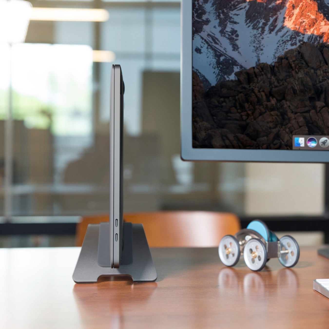 Side view of the Twelve South BookArc MacBook Stand in space grey hold a MacBook upright on a wooden table next to a monitor